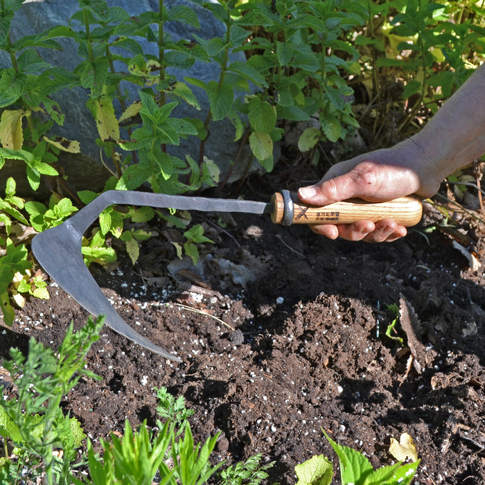 Hand using a hand-forged large weeding hoe with a curved dark steel blade and wooden handle to loosen soil and remove weeds in a sunlit garden bed.