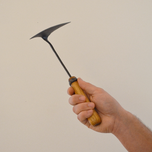 Hand holding a hand-forged winged garden hoe with a dark steel head and wooden handle, shown against a clean neutral background to highlight its size and artisan design.