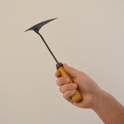 Hand holding a hand-forged winged garden hoe with a dark steel head and wooden handle, shown against a clean neutral background to highlight its size and artisan design.
