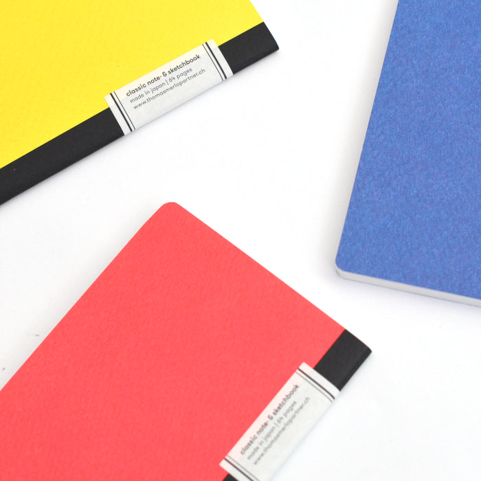 Close-up of colorful pocket notebooks in yellow, red, and blue with black spines, featuring minimalist Japanese design and soft textured covers on a white background.