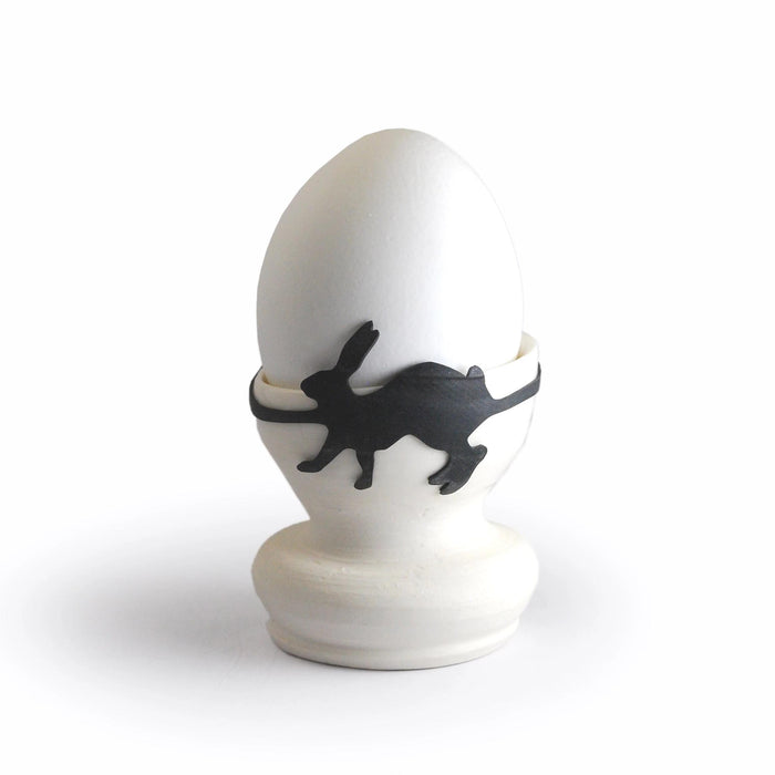 White egg in an egg cup secured with a black rabbit-shaped rubber band, playful animal kitchen accessory and creative Easter table decoration.