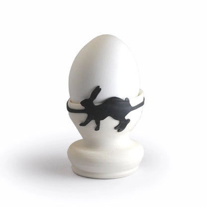 White egg in an egg cup secured with a black rabbit-shaped rubber band, playful animal kitchen accessory and creative Easter table decoration.