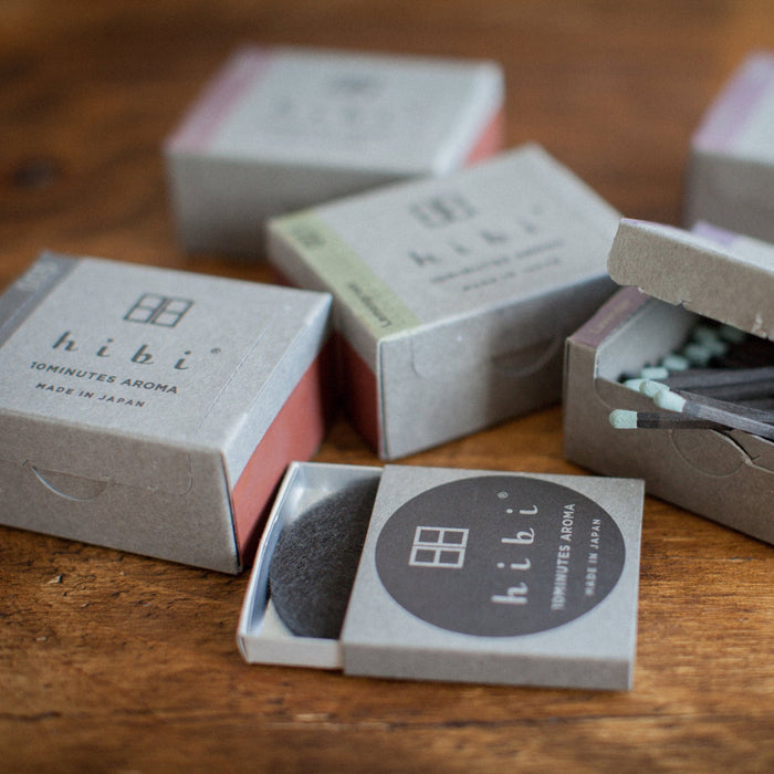 Several hibi 10 Minutes Aroma incense match boxes arranged on a wooden surface, with some boxes open to reveal pastel-tipped incense matches and a small striker pad, creating a relaxed lifestyle-style product display.