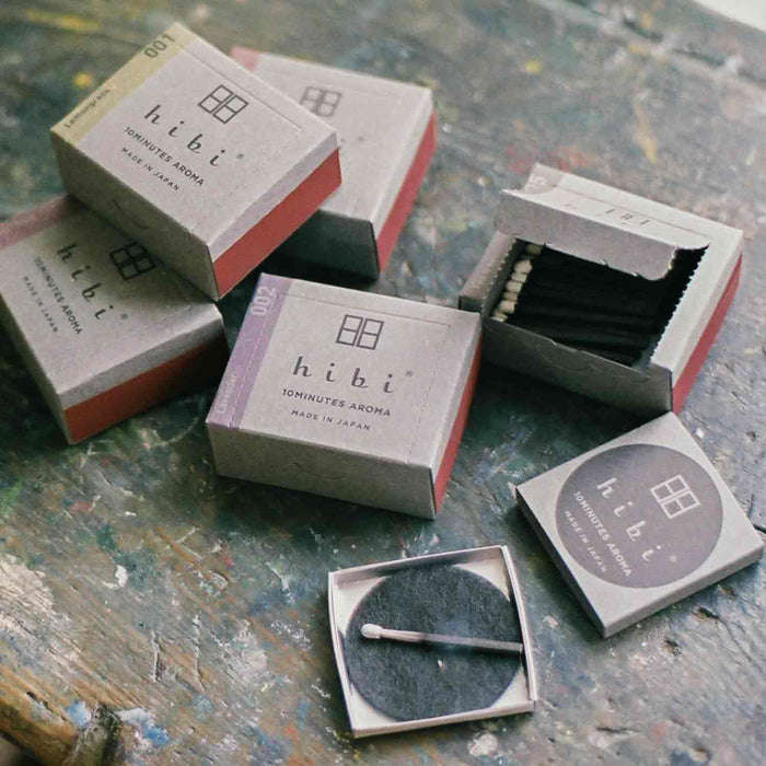 Several hibi 10 Minutes Aroma incense match boxes arranged on a textured, paint-splattered surface, with some boxes closed and others open to reveal black incense matches and a round striker pad, creating an informal lifestyle-style product scene.