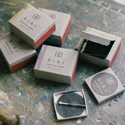 Several hibi 10 Minutes Aroma incense match boxes arranged on a textured, paint-splattered surface, with some boxes closed and others open to reveal black incense matches and a round striker pad, creating an informal lifestyle-style product scene.