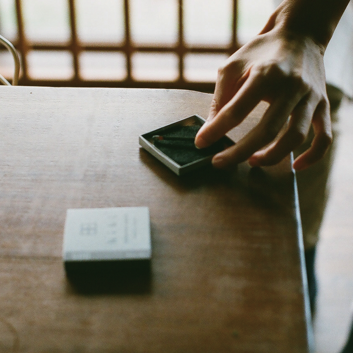 Hand placing a lit hibi incense match on a ceramic striker mat on a wooden table, with a hibi 10 Minutes Aroma incense matchbox in the background.