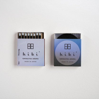 Two Hibi matchboxes side by side on a plain white background. The left matchbox is open, showing the dark match heads and matches inside, while the right matchbox is closed with a blue gradient label.