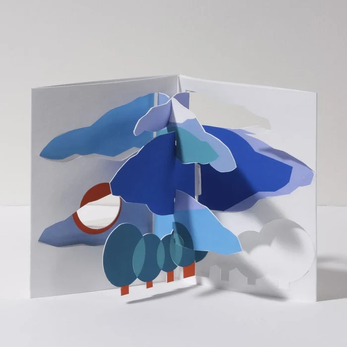 Open 3D pop-up greeting card featuring a stylized blue tree with layered foliage, rounded green trees, clouds, and a rising sun in a modern abstract paper art landscape design.