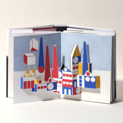 Open art book featuring a colorful 3D pop-up cityscape with geometric buildings in red, blue, yellow, and white, showcasing playful architectural paper art in a modern abstract style.