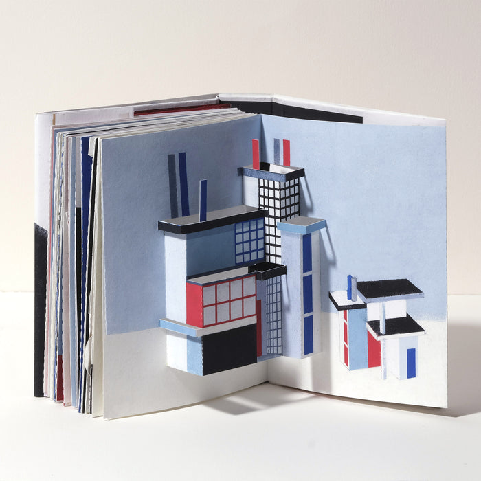 Open art book featuring a geometric 3D pop-up architectural scene with modern buildings in blue, red, black, and white, showcasing abstract paper construction and contemporary design.