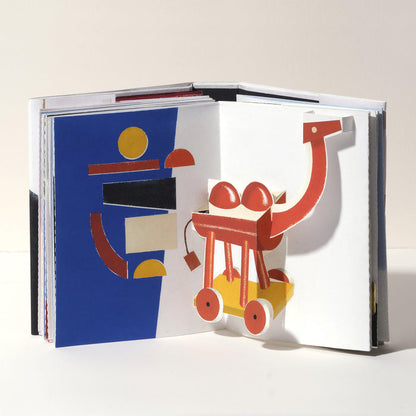 Open art book featuring a whimsical 3D pop-up camel in red, yellow, and white with wheels, paired with bold geometric abstract artwork in blue, red, black, and yellow.