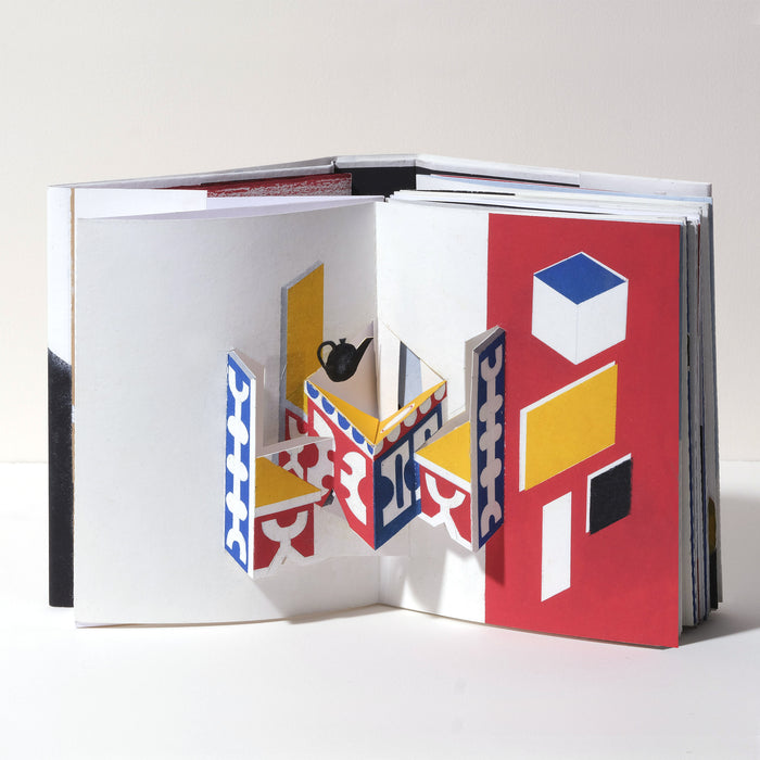 Open art book featuring a geometric 3D pop-up interior scene with abstract table and chairs in red, blue, yellow, black, and white, showcasing modern paper art and contemporary design.