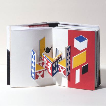 Open art book featuring a geometric 3D pop-up interior scene with abstract table and chairs in red, blue, yellow, black, and white, showcasing modern paper art and contemporary design.