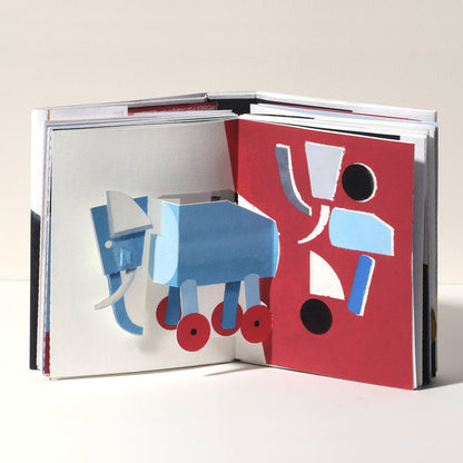 Open art book featuring a playful 3D pop-up blue elephant with wheels, paired with bold geometric abstract artwork in red, black, white, and blue on the opposite page.