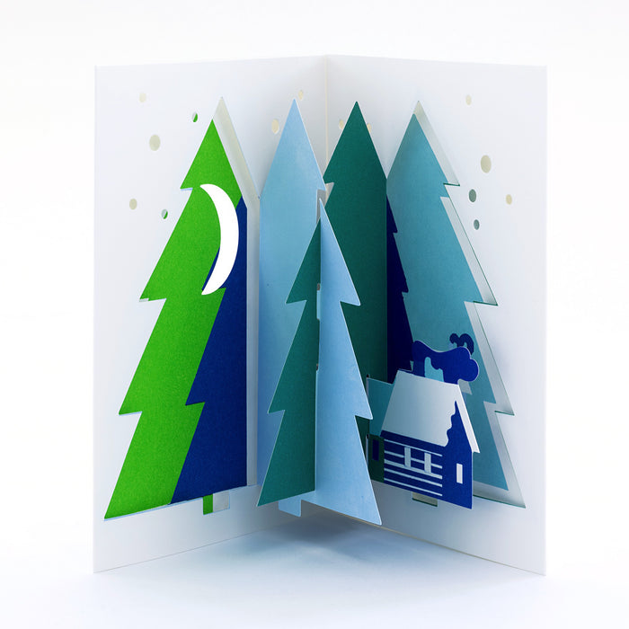 Open 3D pop-up Christmas card featuring a winter forest of layered evergreen trees with a snowy cabin, moon, and falling snow in a minimalist festive paper art design.