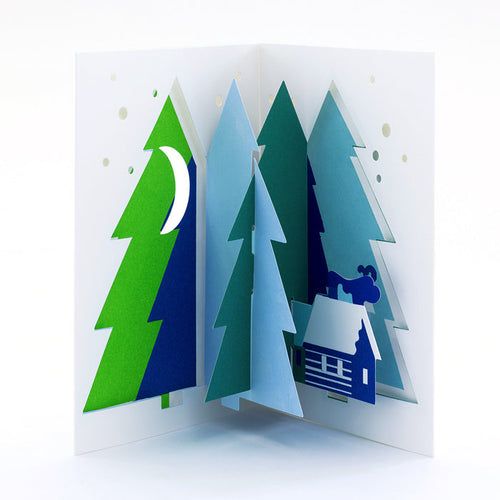 Open 3D pop-up Christmas card featuring a winter forest of layered evergreen trees with a snowy cabin, moon, and falling snow in a minimalist festive paper art design.