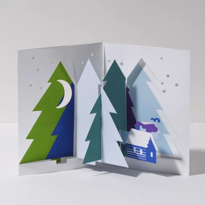 Open 3D pop-up Christmas card featuring a snowy winter forest with layered evergreen trees, a crescent moon, falling snow, and a cozy cabin in a minimalist festive paper art design.