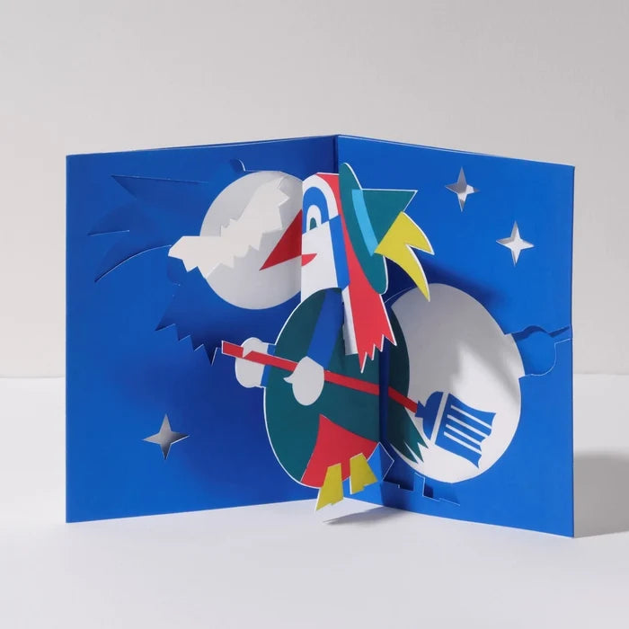 Open 3D pop-up Halloween card featuring a colorful witch flying on a broomstick against a deep blue night sky with stars and a full moon in a playful paper art design.