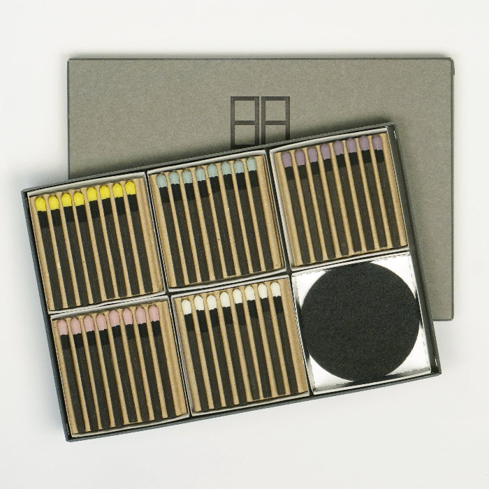Open Hibi large gift box displayed on a white background, showing multiple compartments of incense matches in assorted colors alongside a round black burning mat, with the matching gray box lid placed behind.