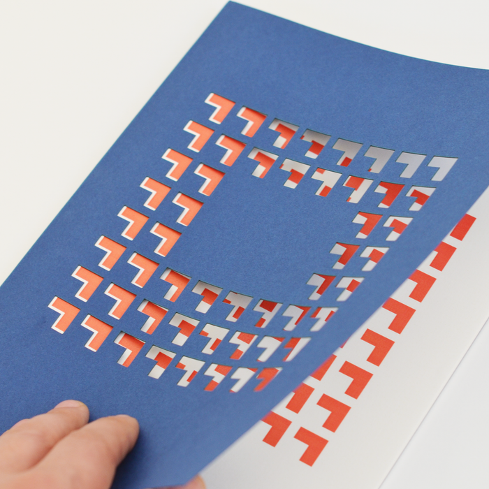 Hand holding a blue TÄT-TAT designer notebook with geometric die-cut cover revealing red pattern underneath, modern creative notebook for writing and sketching.