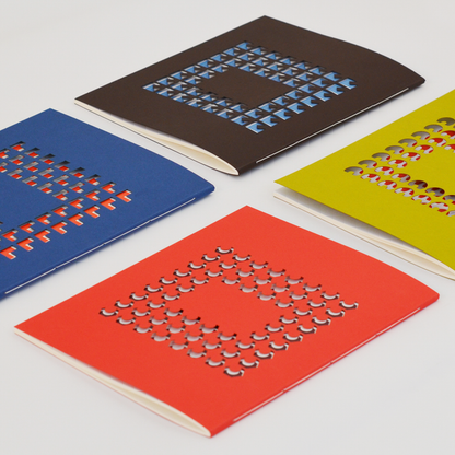 Colorful TÄT-TAT designer notebooks with geometric die-cut covers in red, blue, yellow, and brown, modern creative notebooks for writing, sketching, and journaling.