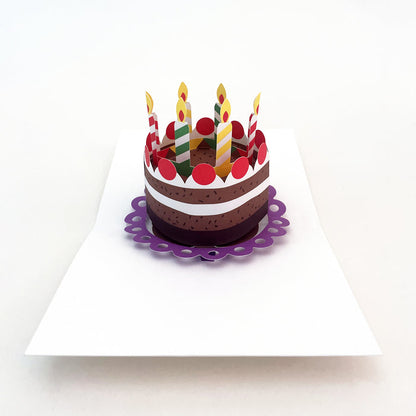 Open 3D pop-up birthday card featuring a layered cake with colorful striped candles and red decorations on a white card base, festive paper art greeting card for birthdays and celebrations.