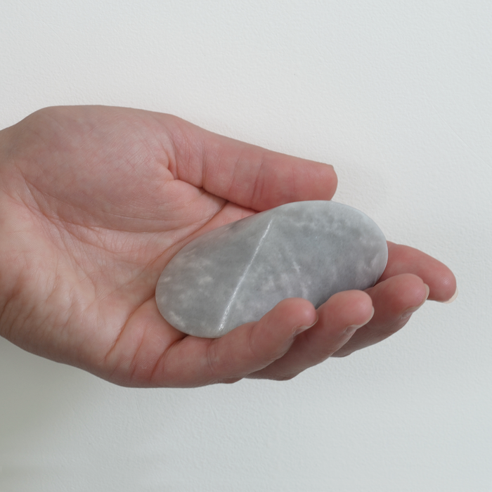 Hand holding a smooth light gray marble oloid sculpture, shown against a clean white background to highlight its size, shape, and natural stone texture.