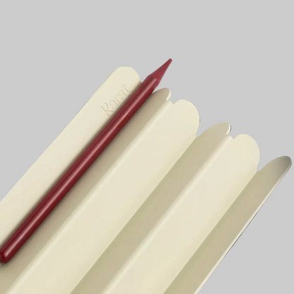 Close-up of a cream-colored ridged pen tray with a red pencil resting in one groove, featuring rounded edges and a subtle embossed logo, photographed on a light gray background.
