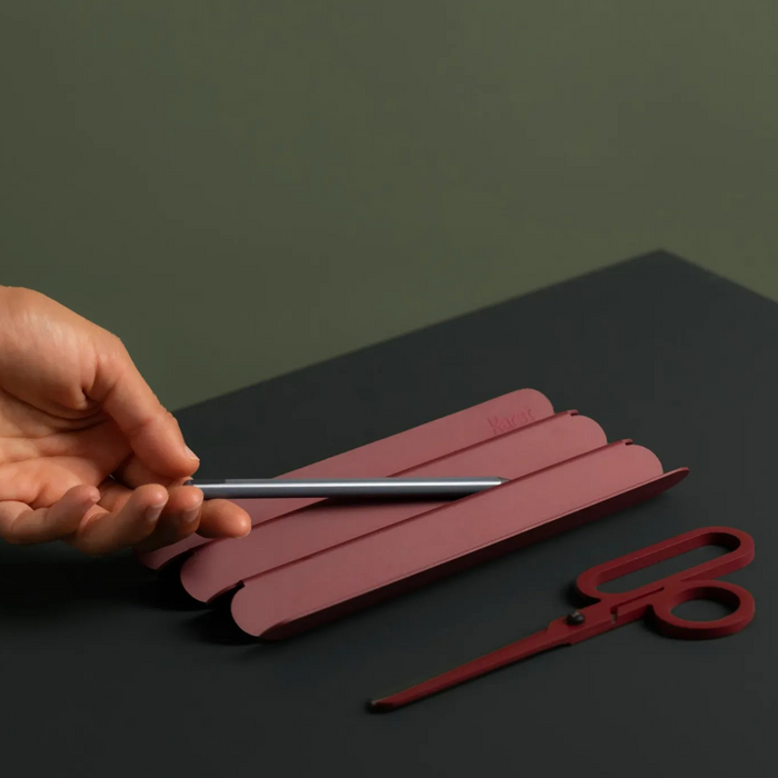 Hand placing a silver pen onto a burgundy, ridged pen tray on a dark tabletop, with matching red scissors beside it, styled in a minimalist workspace setting.