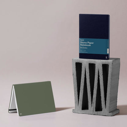 Two Karst stone paper notebooks displayed in a minimalist studio setting: a navy Pro Series notebook standing upright on a concrete block and an olive green notebook folded tent-style on the surface beside it, against a soft neutral background.
