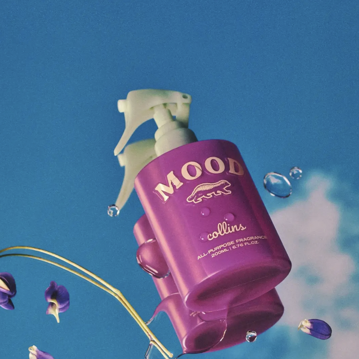 A purple room spray bottle with a label that reads 'MOOD collins'. The background features a serene sky with clouds and floating violet flowers.