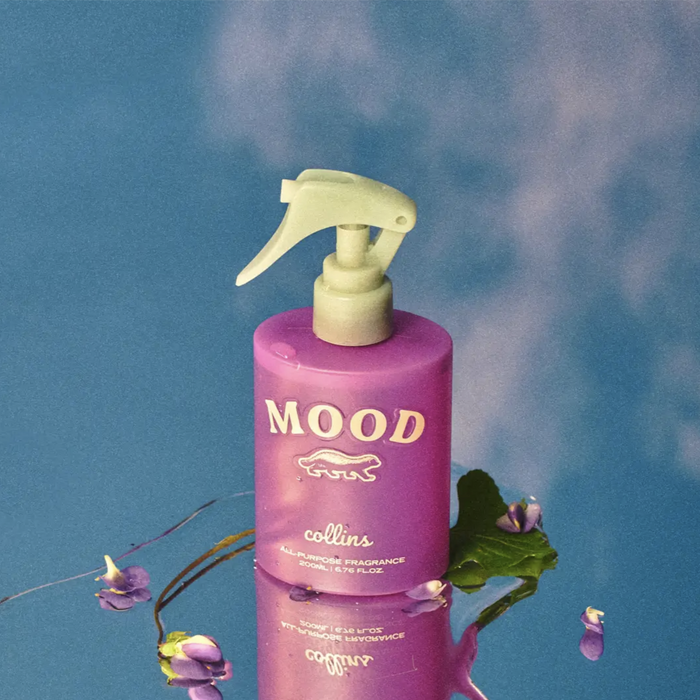 Mood all-purpose fragrance spray bottle in a purple finish, styled with violet petals on a glossy blue reflective surface for a fresh floral home scent product display.