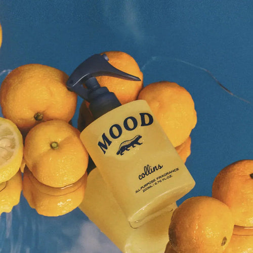 A clear bottle of 'MOOD' room spray with a pump dispenser, surrounded by lemons, against a blue background.