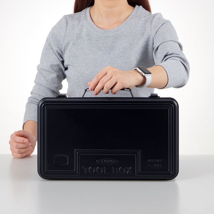 Person wearing a gray long-sleeve shirt holding a black rectangular toolbox with a handle, placed on a white table against a white background.